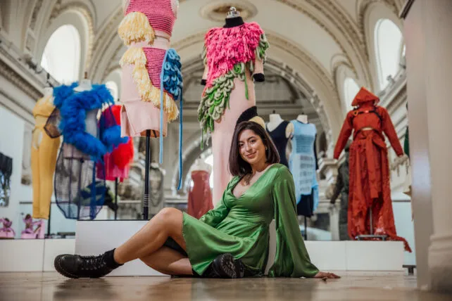 A young woman with dark hair and a light complexion sits on the floor in a bright, spacious room with an arched ceiling and tall windows in the background. She is wearing a flowing, long-sleeved green satin dress and chunky black boots, looking towards the camera with a slight smile. Behind her, several avant-garde fashion garments are displayed on mannequins, each featuring unique textures, colors, and designs. These include a pink top with yellow and blue woven elements, another pink garment with vibrant pink and green feathery textures, a blue feathery piece, a yellow woven garment, and a dramatic red hooded outfit. The overall setting appears to be an art gallery or a fashion exhibition.