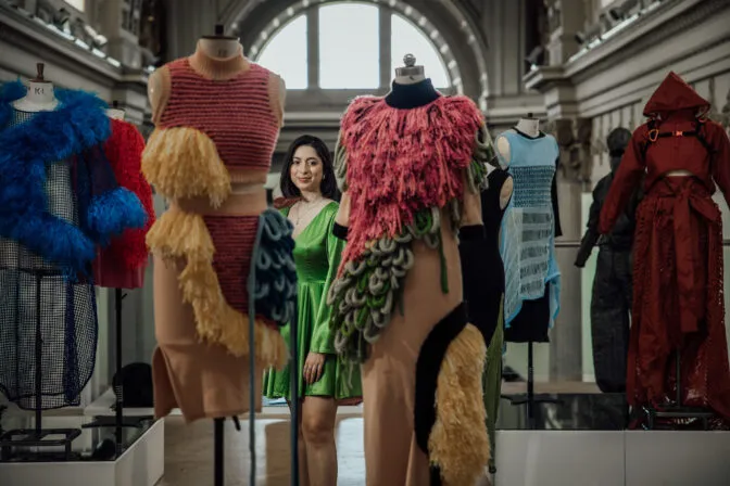 A young woman with dark hair and a light complexion stands in the middle of a fashion exhibition, looking directly at the camera with a subtle smile. She is wearing a green satin dress. She is surrounded by several mannequins displaying avant-garde garments. In the foreground, a mannequin on the right wears a pink garment adorned with vibrant pink fluffy textures and green, leaf-like woven elements. Another mannequin in the foreground on the left features a striped red and pink knit top with a yellow textured overlay. In the background, other unique pieces are visible, including a blue feathery garment, a red hooded piece with straps, and other textured designs. The setting appears to be a gallery with high ceilings and large arched windows in the background.