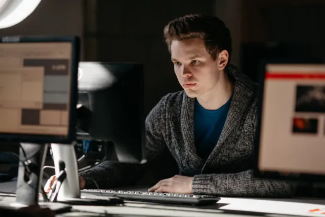 A young man with light brown hair and fair skin sits intently in front of a computer, looking at the screen. He is wearing a blue t-shirt under a grey, textured cardigan. His left hand rests on the computer mouse, and his right hand is on the keyboard. The lighting is somewhat dim, with light primarily falling on his face and the monitors. There are two other blurred computer monitors in the foreground, one on the left and one on the right, showing various interfaces.